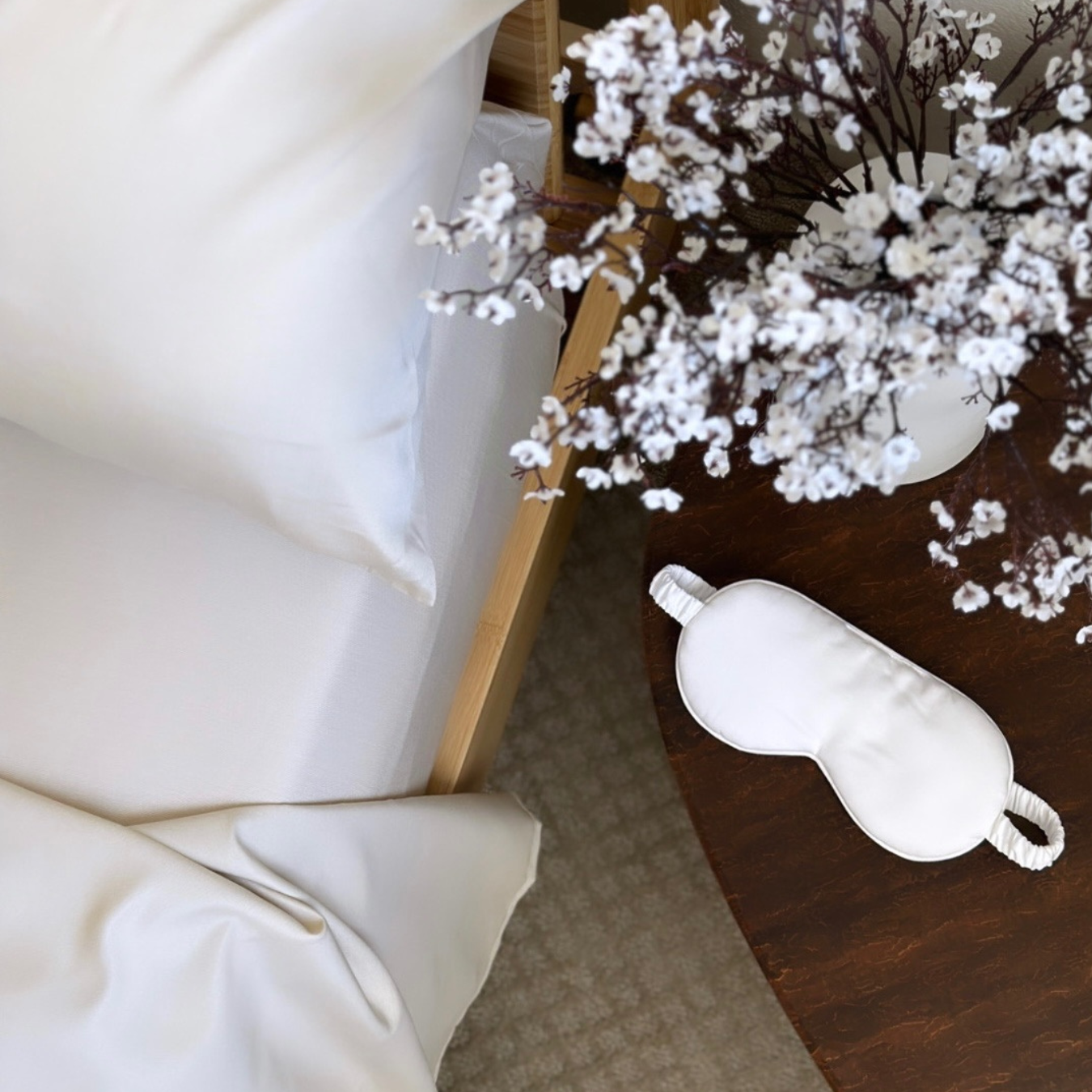 White Copper Infused Bamboo Pillowcase Set on a wooden bed with delicate white flowers and a silk eye mask.
